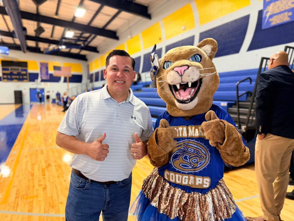 Commissioner Gus Ruiz stands with the Mascot of Santa Maria smiling at the camera.